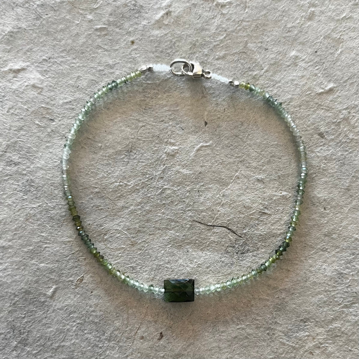 Forest green tourmaline bracelet with tiny beads and darker rectangular center stone on textured surface