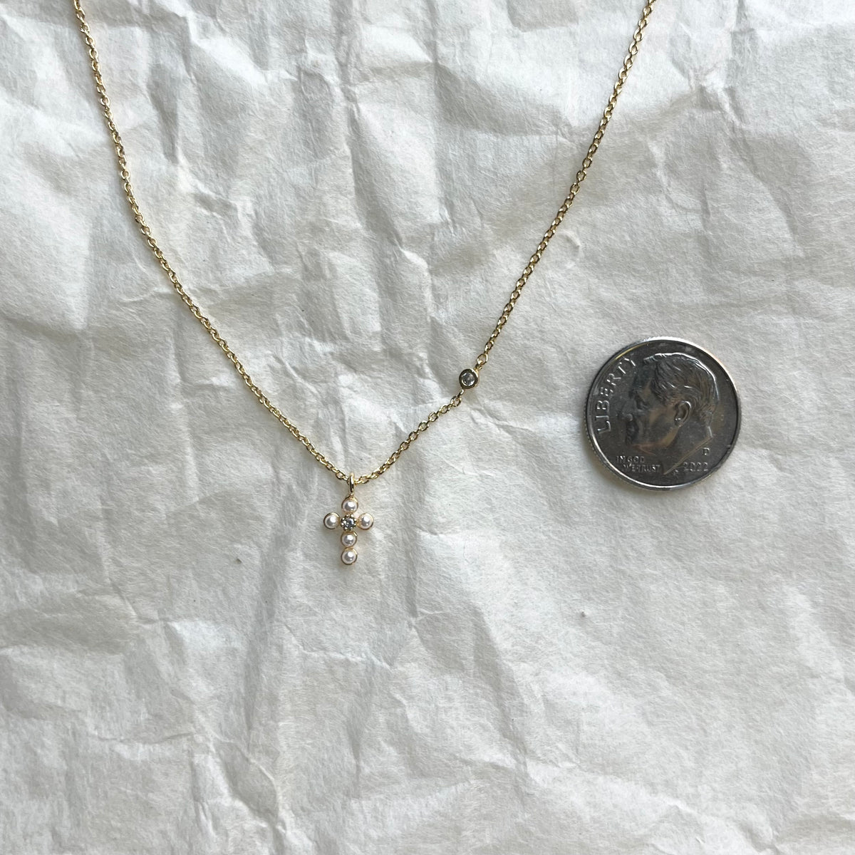 Delicate gold chain necklace with small pearl cross pendant beside dime for scale
