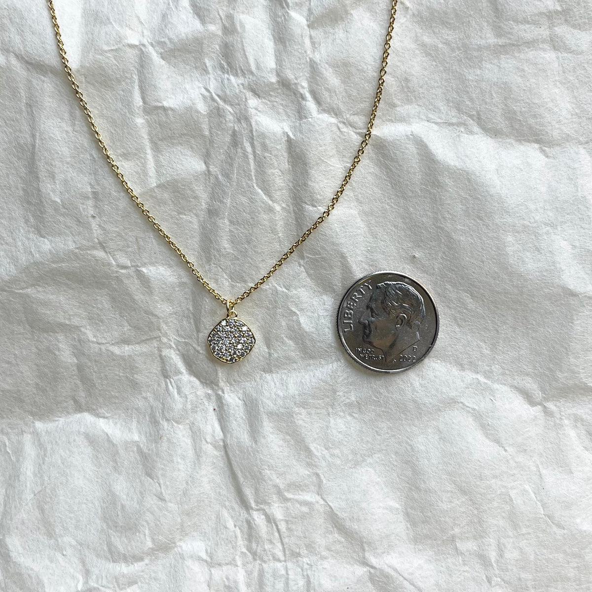 Gold CZ disc necklace on white fabric next to dime for size comparison