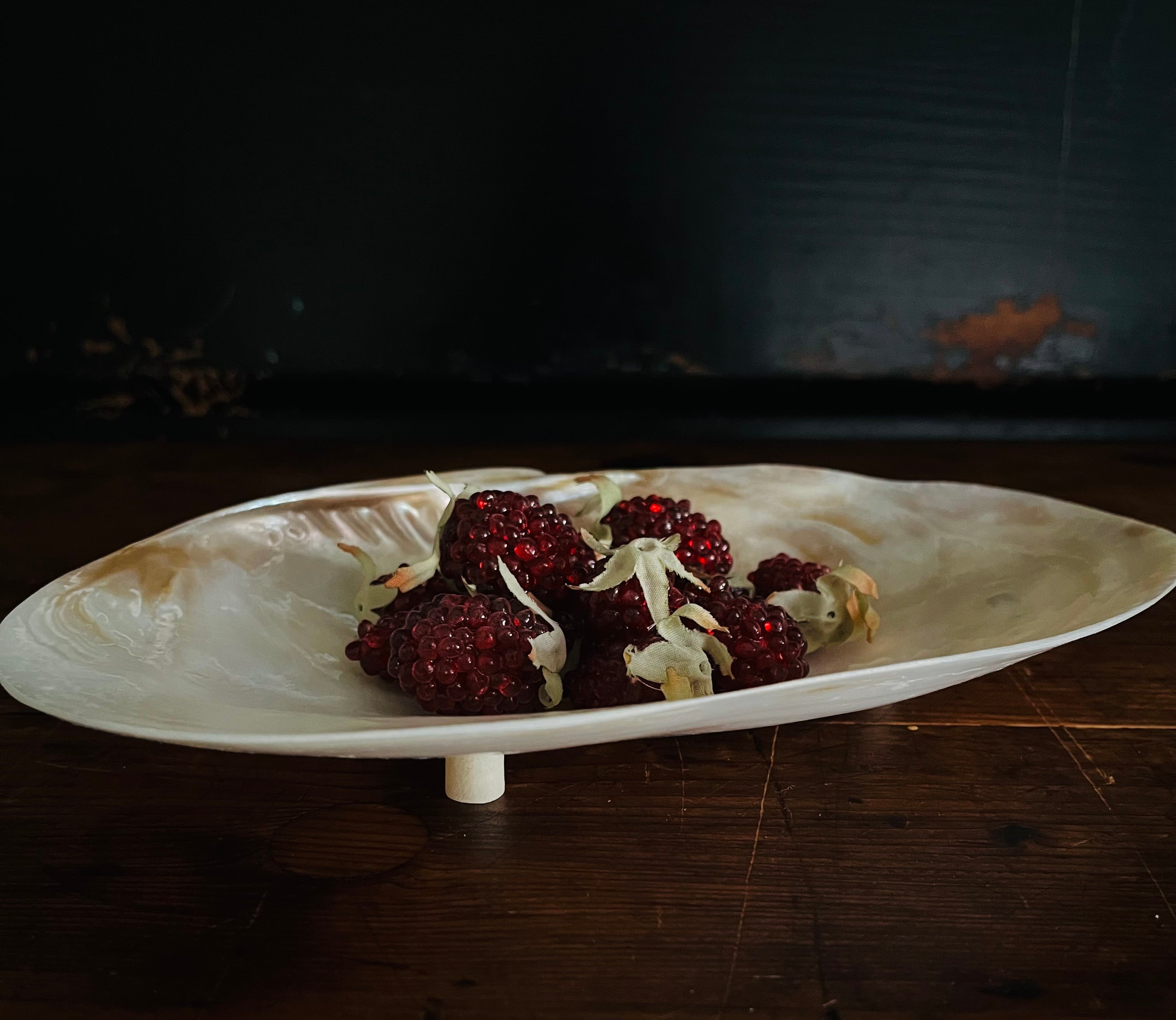 White oval seashell footed dish on wooden table holding dark berries and petals