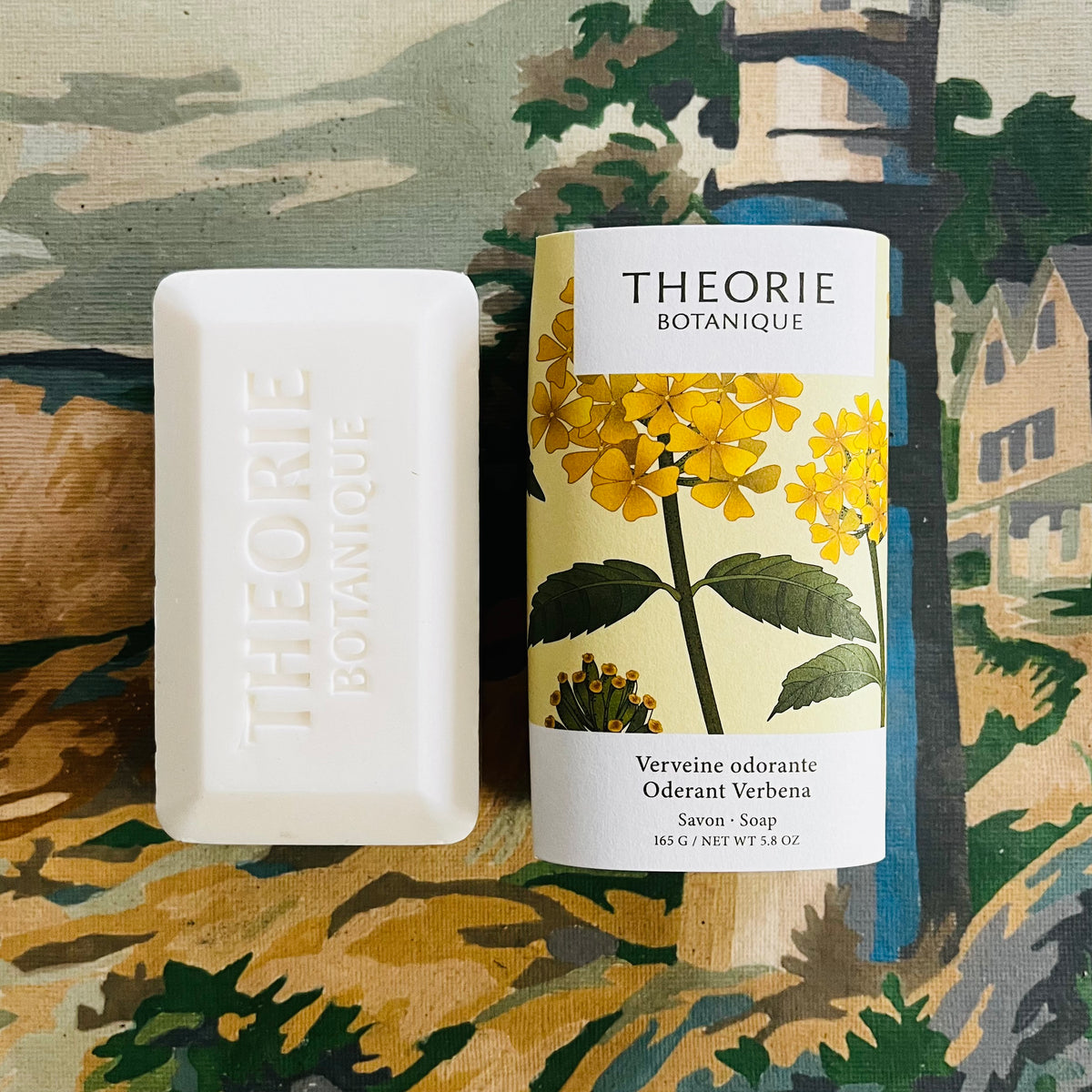 White Theorie Botanique bar soap beside yellow floral packaging on painted fabric background