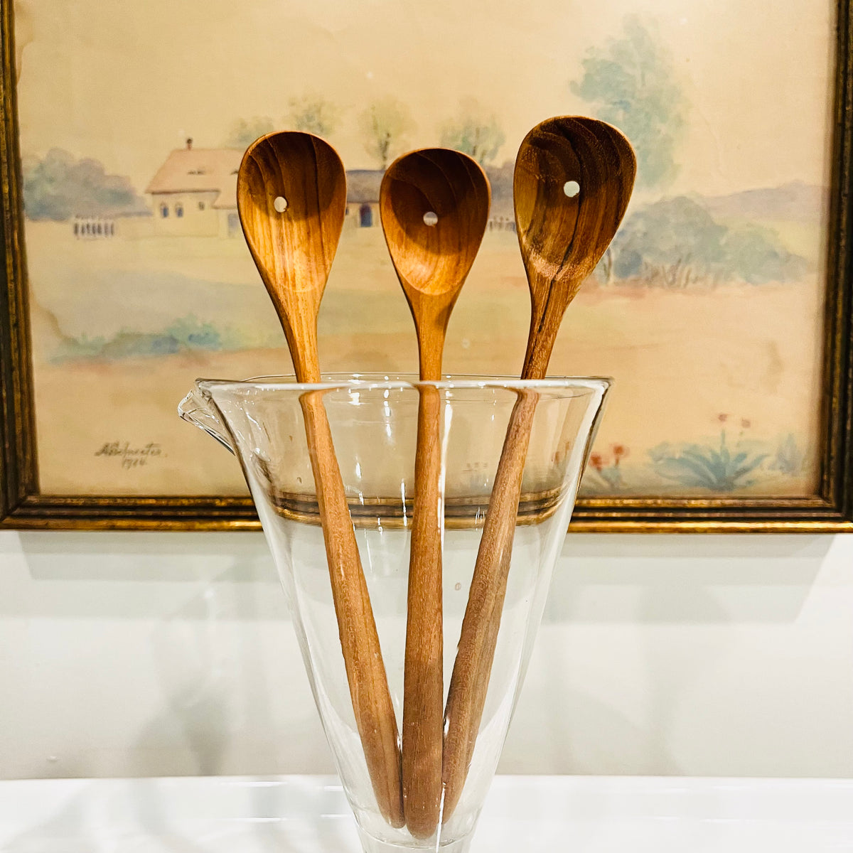 Three teak olive spoons with hanging holes displayed in a clear glass pitcher