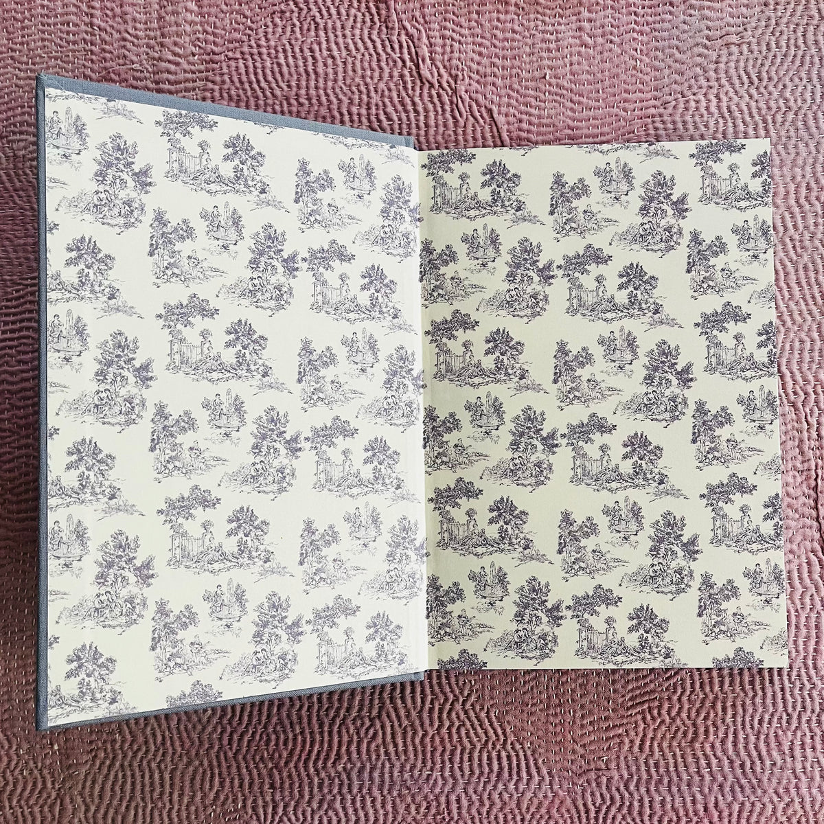 Open journal with cream pages featuring repeating black toile pastoral pattern on pink woven surface