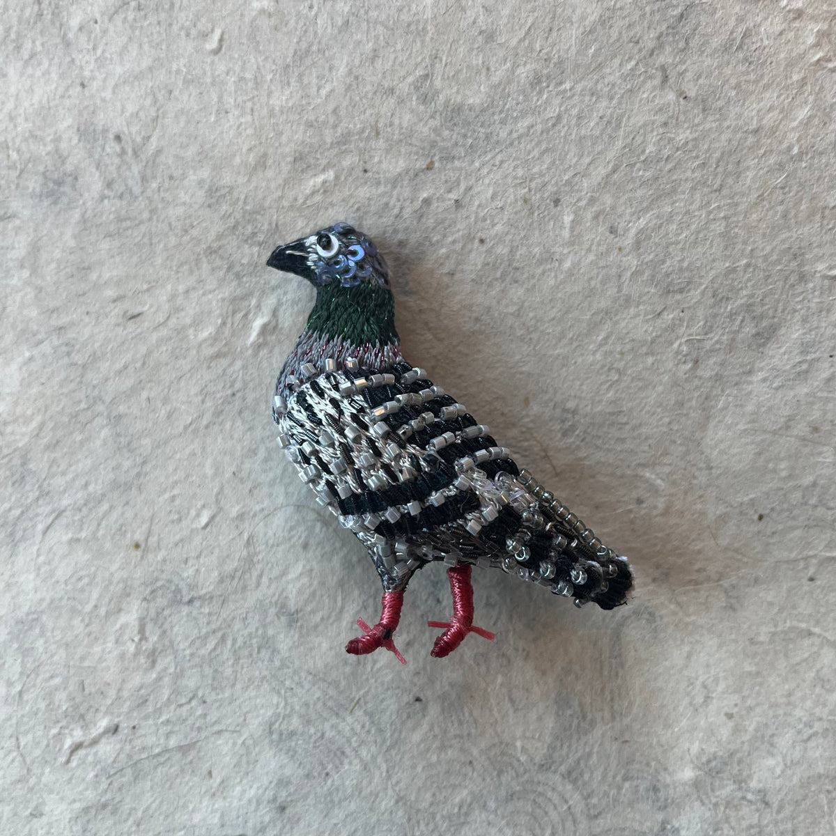Beaded pigeon brooch with iridescent neck, barred wings, and red legs on textured background