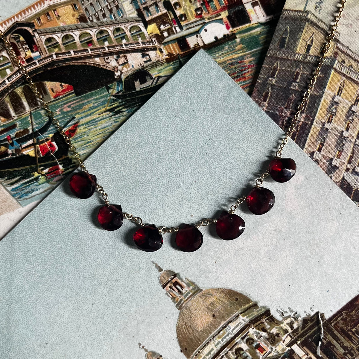 Way to Your Heart Necklace with deep red disc charms on gold chain, displayed on Venice postcard