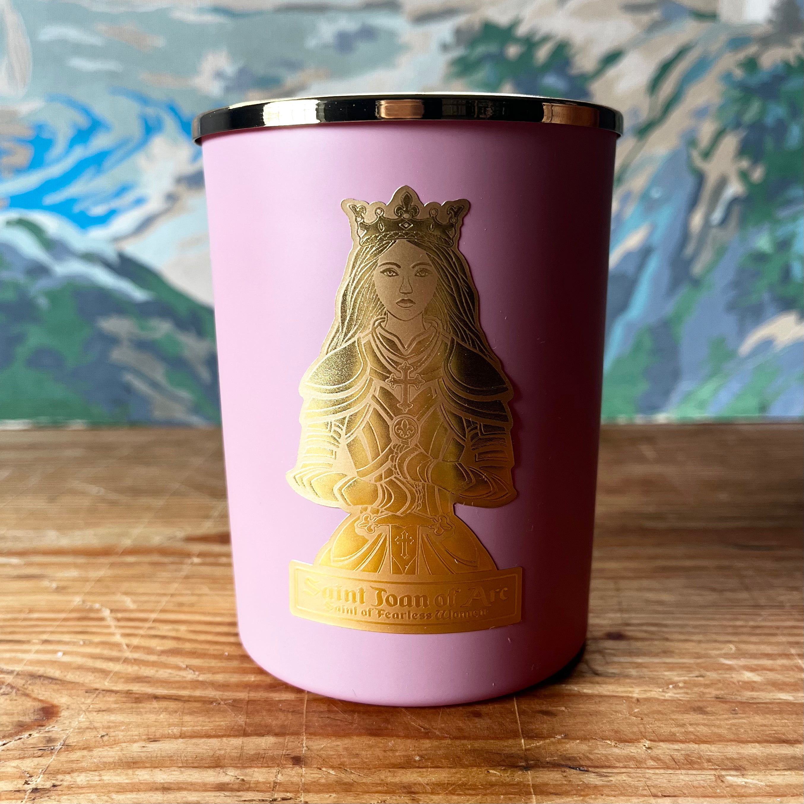 Pink tumbler candle with gold Saint Joan of Arc emblem on wooden surface