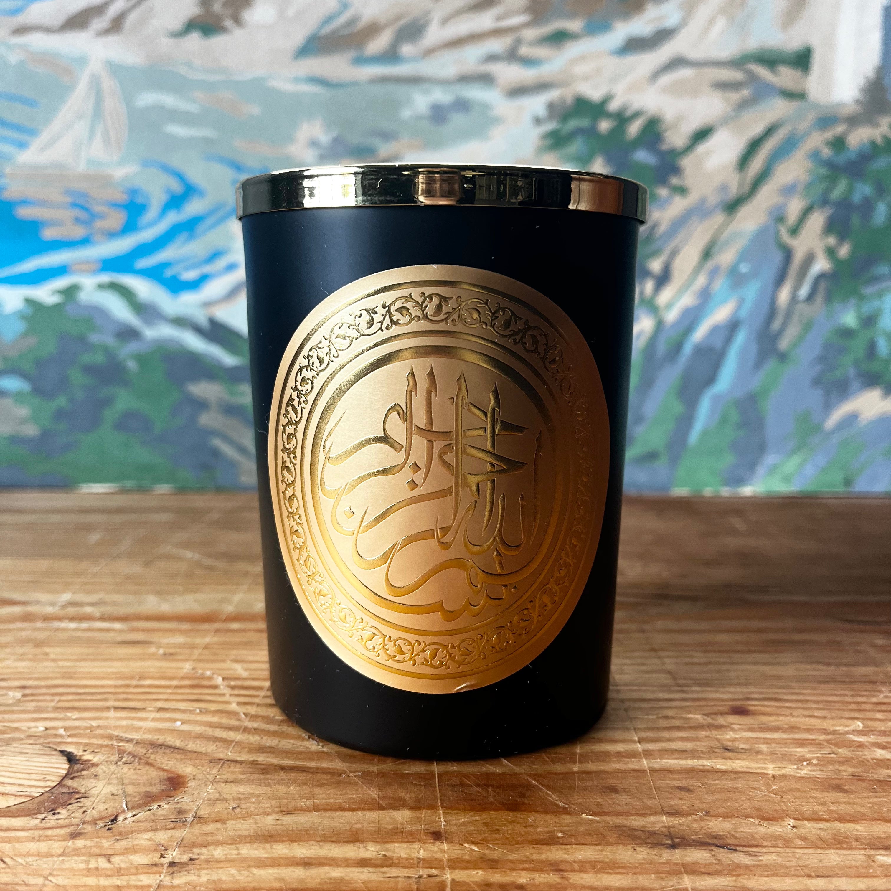 Black glass candle with gold bismillah medallion on wooden table with painted backdrop