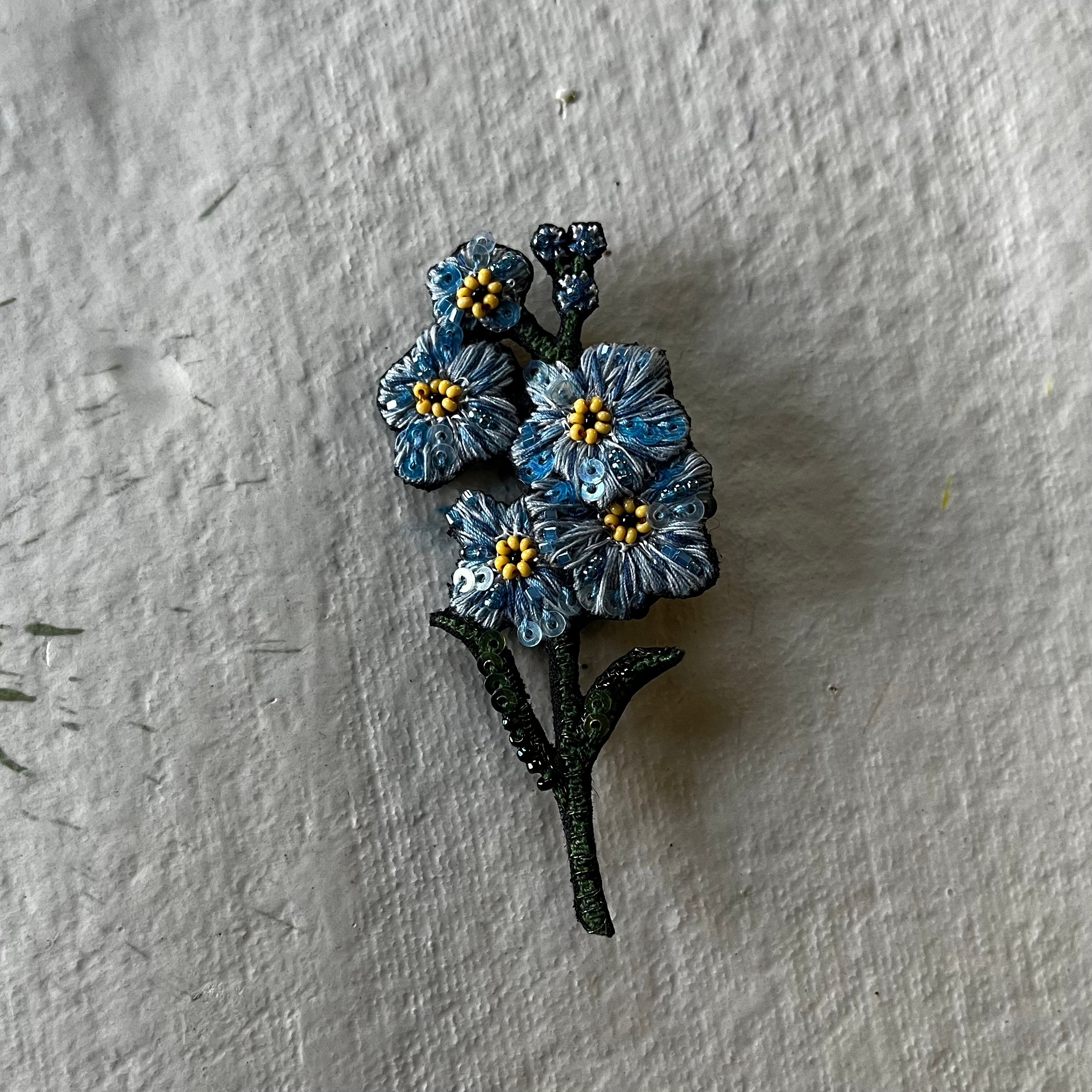 Blue forget-me-not brooch with yellow centers and dark green stem on white textured background