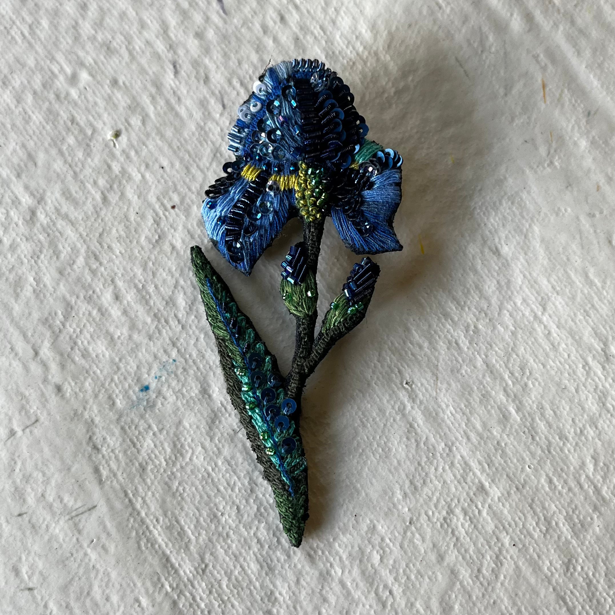 Wild iris brooch with blue sequined petals and green beaded leaves on white background