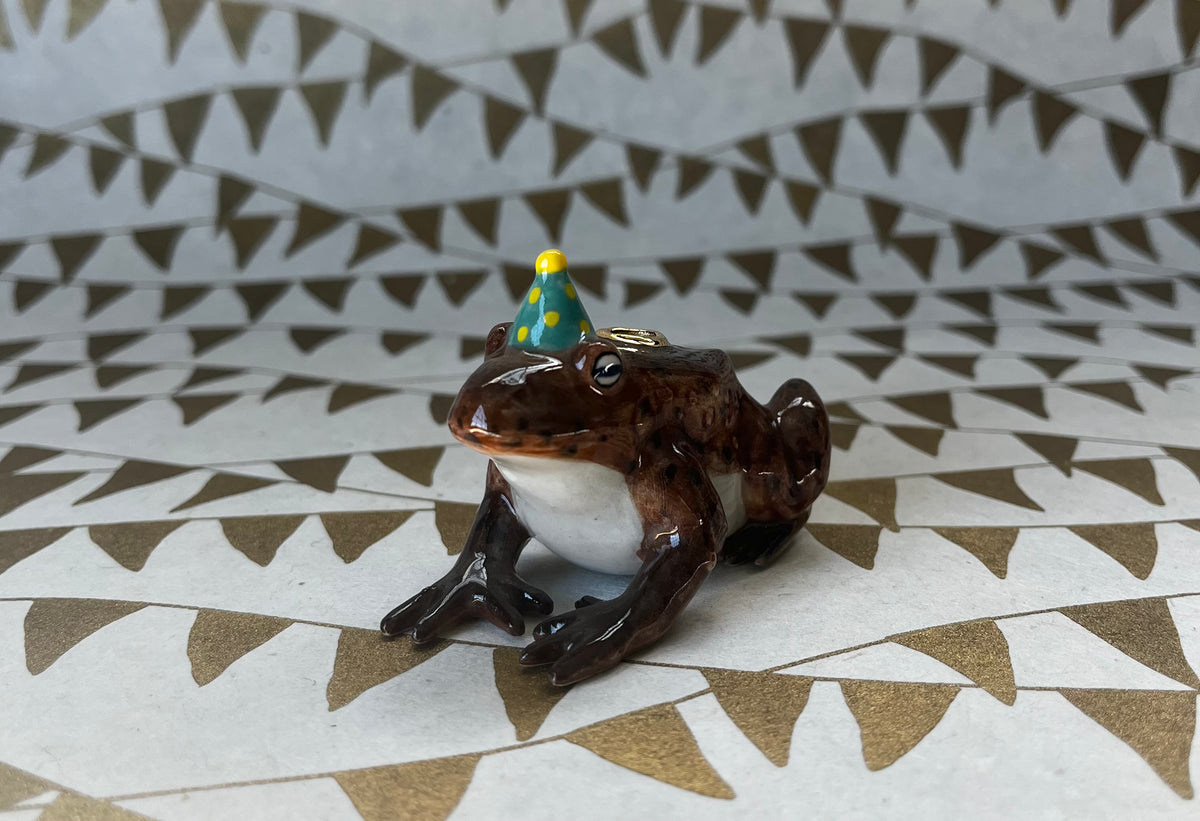 Brown ceramic toad figurine with teal and yellow party hat on patterned surface