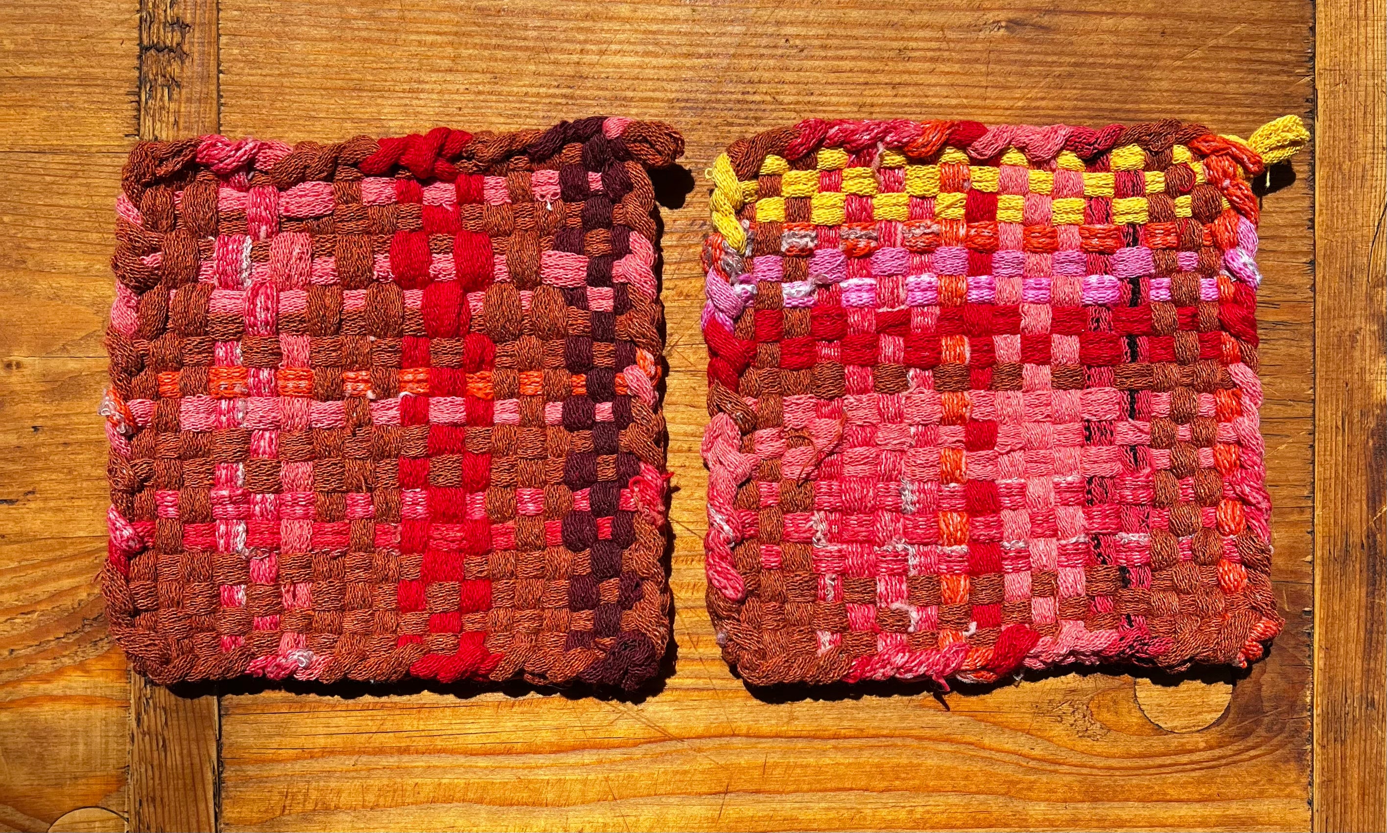 Two woven potholders in shades of red, pink, brown, and yellow on wood surface