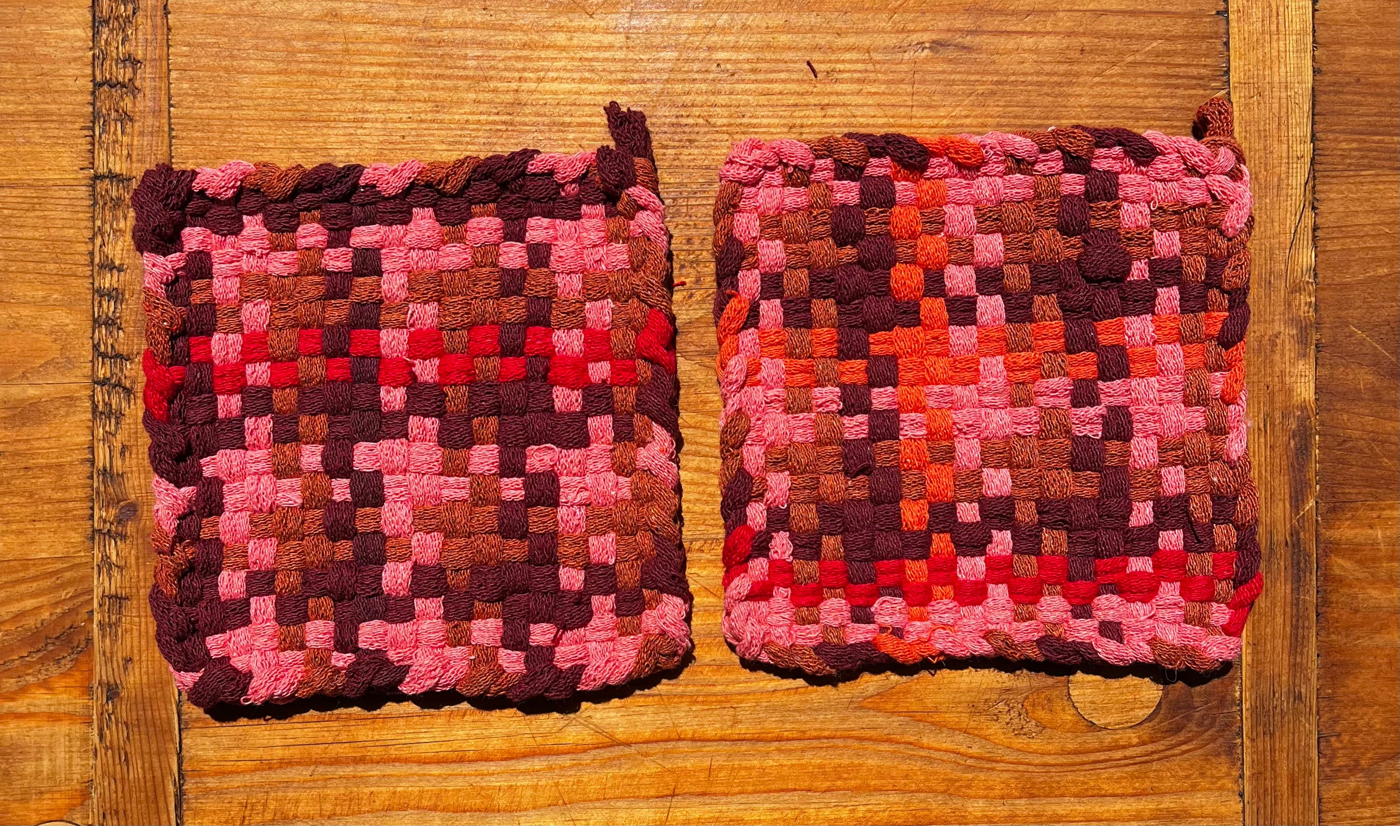 Two woven Nuts & Fruits potholders in pink, burgundy, and red checkered pattern on wooden surface