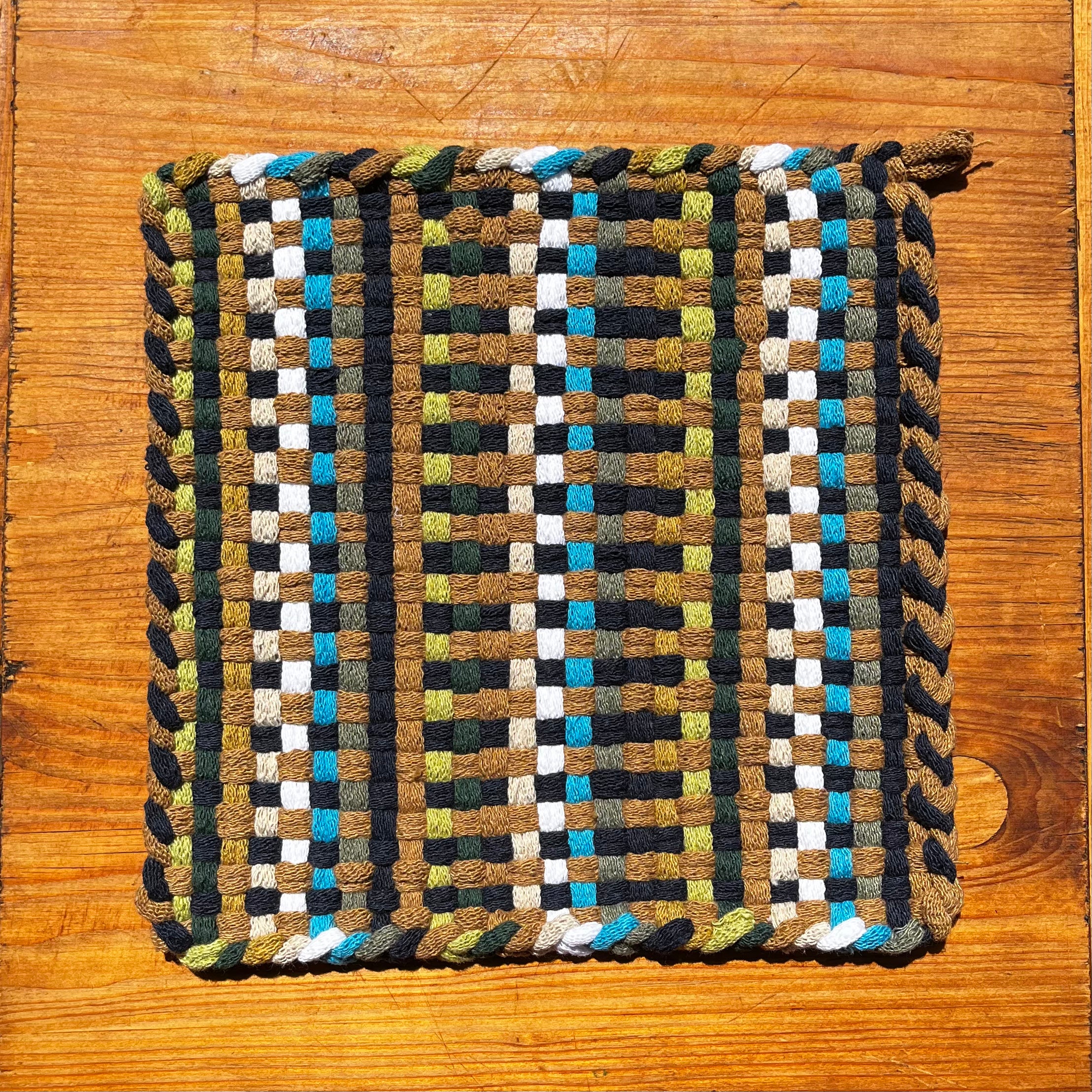Woven Seaside Potholder with checkered pattern in blues, greens, browns, and black on wooden surface