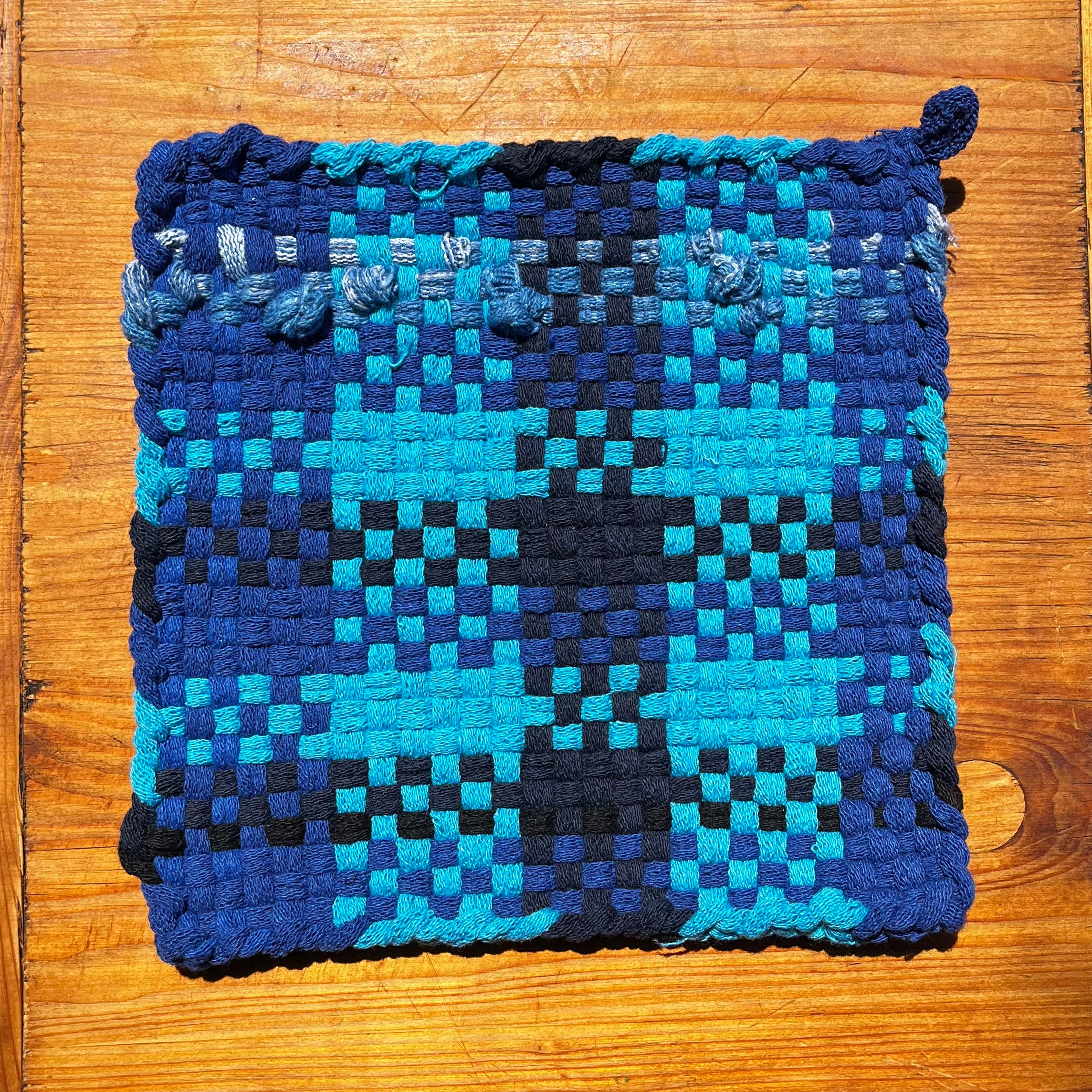 Woven potholder in blue, black, and turquoise checkered pattern on wooden surface.