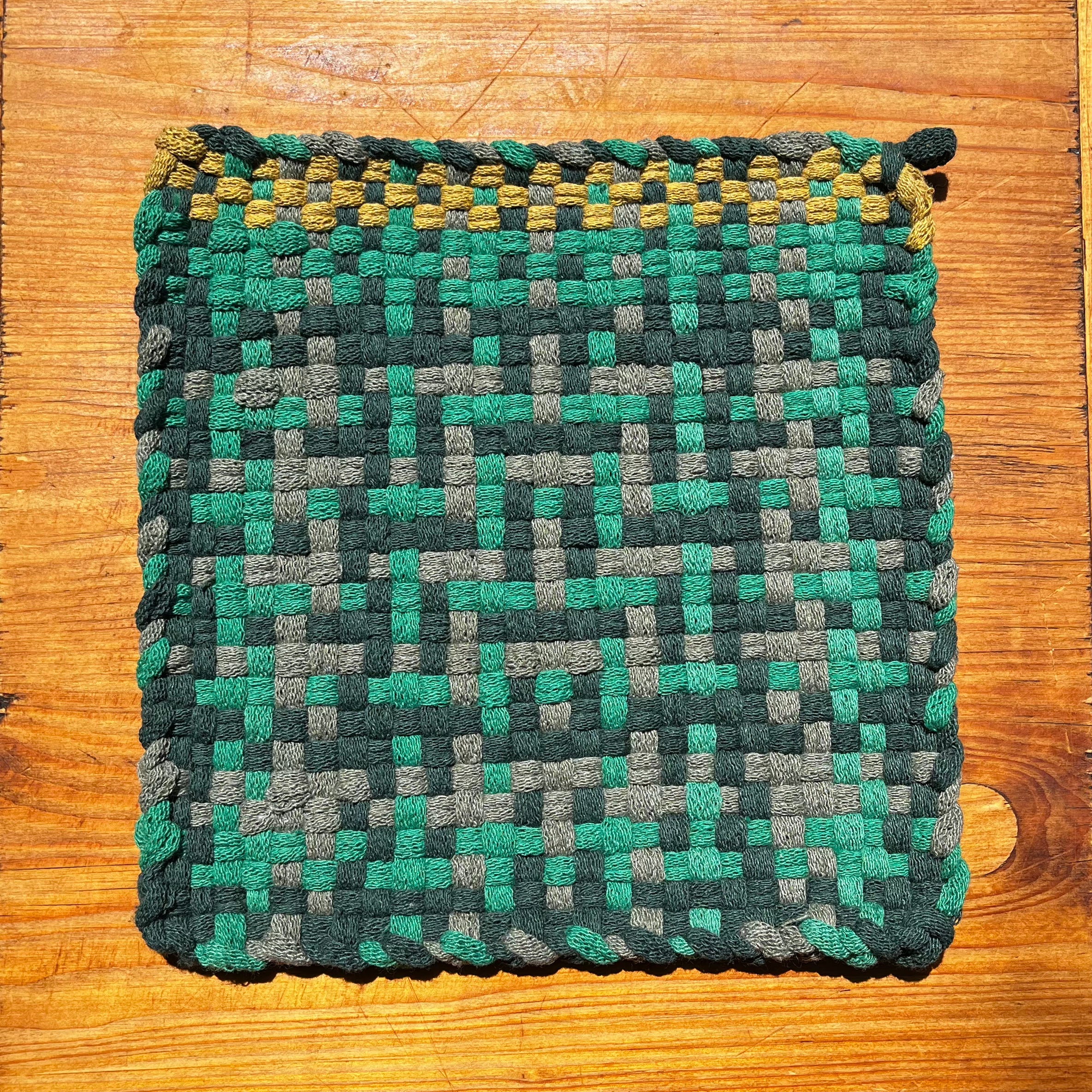 Peak into the Jungle Potholder with woven checkerboard pattern in teal, gray, and yellow on wood surface