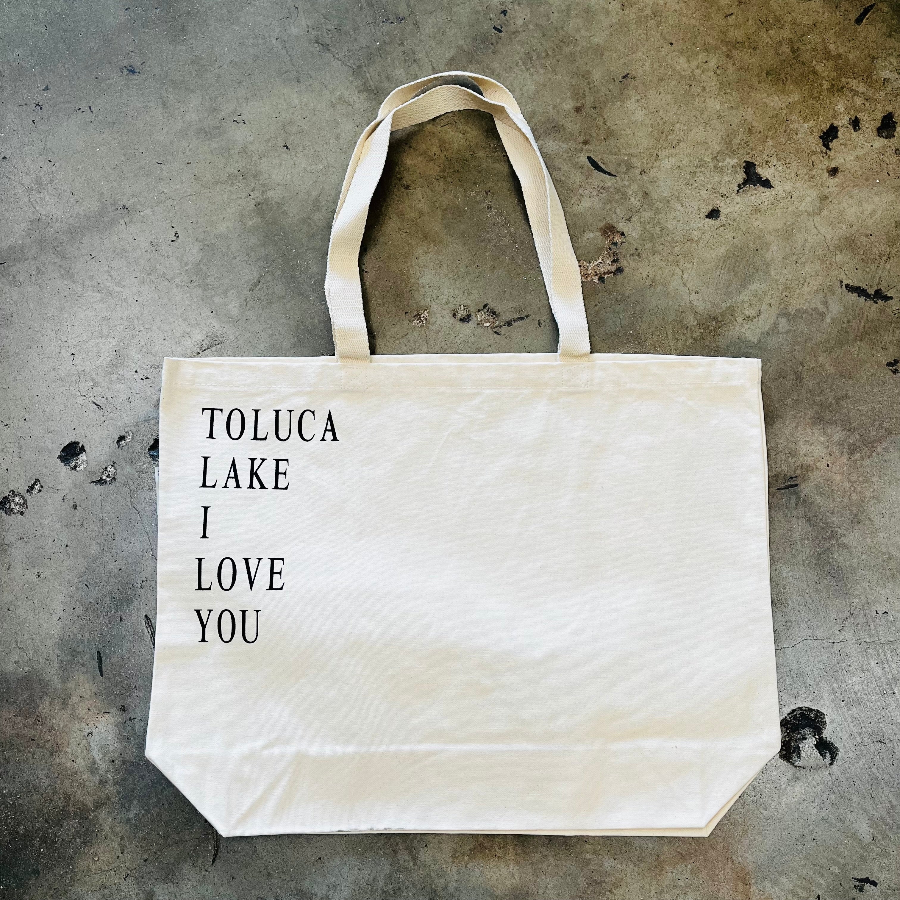 Cream canvas tote bag with black text reading Toluca Lake I Love You on concrete floor