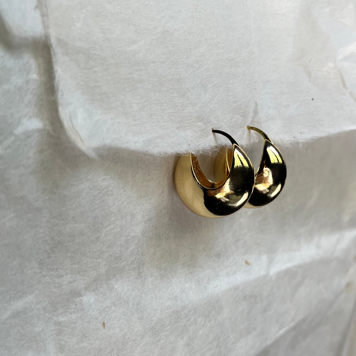 Gold huggie hoop earrings lying on white fabric surface, showing curved thick design